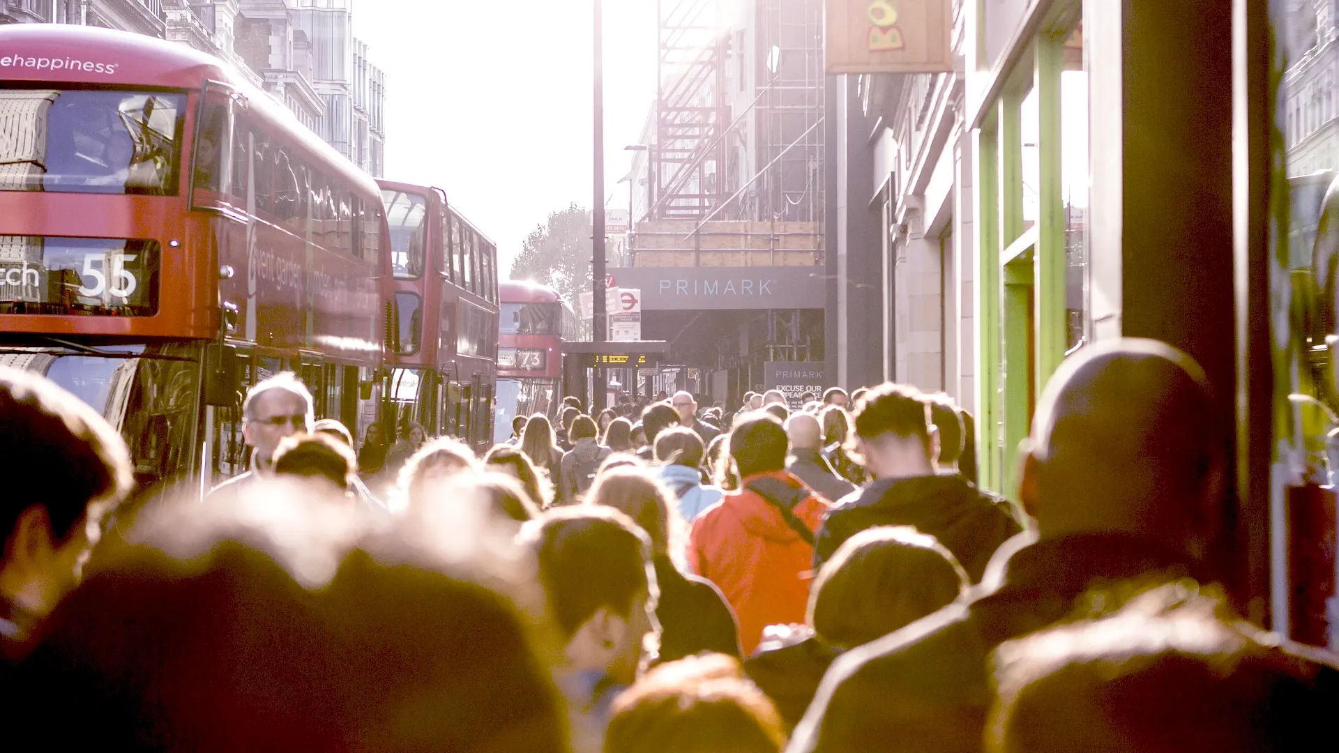 A crowd on a busy London street with three red buses picking up passengers to the side